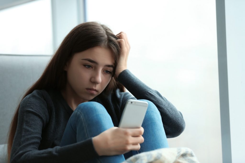 Upset teenage girl with smartphone sitting at window indoors.